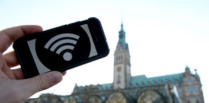 Auf einem Handy ist das Wlan-Symbol weiß auf schwarz zu sehen, dahinter das Hamburger Rathaus