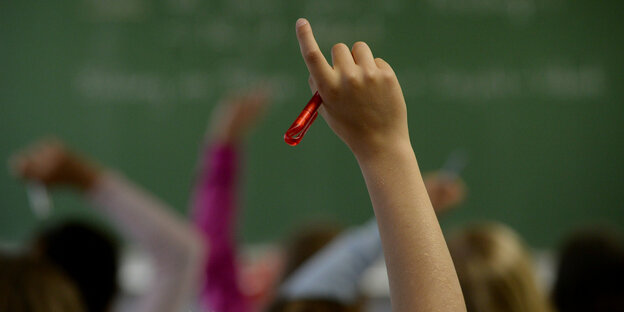 Ein Kind meldet sich im Unterricht. man sieht nur den nackten Unterarm und die Hand, die gleichzeitig einen roten Stift hält. Im Hintergrund andere Kinde rund eine grüne Tafel.