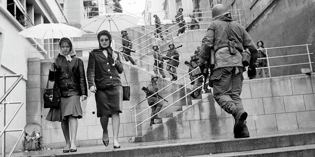 Zwei Frauen mit Regenschirmen gehen eine Treppe hinunter, hinauf gehen Soldaten mit Gewehren